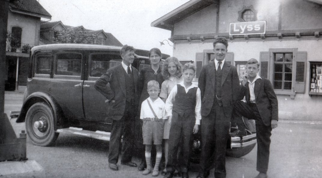 Black and white photograph of a family posing in front of an old car with a building labeled 'Lyss' in the background.